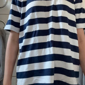 Randig t-shirt i marinblå och vit - Säljer en klassisk t-shirt med breda marinblå och vita ränder. T-shirten har korta ärmar och en avslappnad passform, perfekt för en chill och stilren look. Tillverkad i mjuk bomull som känns skön mot huden.