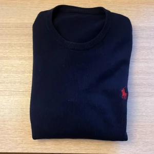 Mörkblå stickad tröja från Ralph Lauren - Klassisk mörkblå stickad tröja från Ralph Lauren med rund halsringning och röd broderad logga på bröstet. Tröjan har långa ärmar och ribbade muddar vid hals, ärmslut och nederkant. Perfekt för en stilren och chill look.