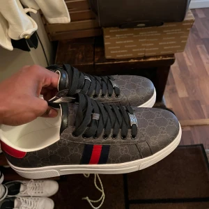 Gucci sneakers med GG-mönster Storlek 43/44 - Snygga Gucci sneakers i mörkgrått med klassiskt GG-mönster, svarta snören och vita sulor. Skorna har röda och marinblå ränder på sidan samt röd häl med Gucci-logga. Obs ( Box samt dust bag tillkommer) 🙌 Hör av er vid minsta lill bekymmer!