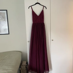 Vinröd långklänning med spetsdetaljer - Säljer en vinröd långklänning med smala axelband och v-ringad urringning. Överdelen har vackra spetsdetaljer med blommönster och en liten knytning i ryggen. Kjolen är i tunn, luftig tyll som ger ett elegant fall. Perfekt för bal eller fest!