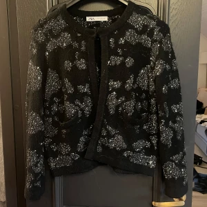 Svart glittrig kofta från Zara - Säljer en svart kofta från Zara i storlek M med glittriga paljettdetaljer över hela plagget. Koftan har rund halsringning, långa ärmar och två små fickor framtill. Perfekt för dig som vill sticka ut med en snygg och trendig look.