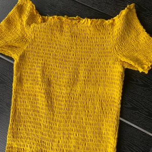 Gul smockad topp från H&M - Superfin gul smockad topp från H&M med korta ärmar. Toppens material är stretchigt och har en rynkad struktur som ger en cool och trendig look. Perfekt för att liva upp din outfit med en färgklick!