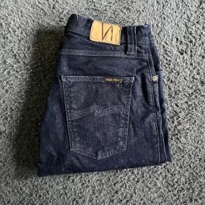 Nudie jeans  - Snygga mörkblå skinny jeans från Nudie med klassisk femficksdesign och läderpatch bak i midjan. Jeansen är i stretchigt denim och har en smal passform som sitter tight längs benen. Perfekta för dig som gillar en stilren och enkel look. Längd 112cm. Ifall det inte finns någon bild i annonsen där jag bär plaggen jag säljer så är det för att jag ej kan ha storleken heller passformen, så kolla måtten för storlek och googla på modellnamnet för passform 😉