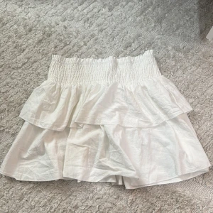 Vit volangkjol från SHEIN - Supersöt vit kortkjol från SHEIN med bred smock i midjan och dubbla volanger. Kjolen har en luftig och flowy känsla, perfekt för att skapa en fräsch och trendig look. Passar dig som gillar ljusa toner och romantiska detaljer.