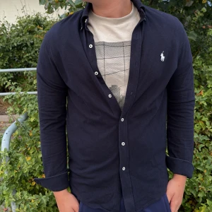 Ralph lauren skjorta - Snygg mörkblå skjorta från Ralph Lauren i lätt mesh-bomull. Klassisk design med knappar hela vägen och ikoniska broderade loggan på bröstet. Perfekt att bära öppen över en t-shirt eller knäppt för en clean look. Ny pris 1600 Inga defekter pris kan diskuteras vid snabb affär 😁