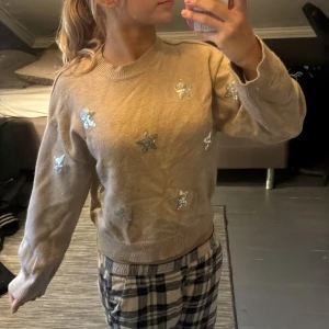 Beige stickad tröja med stjärnor - Supermysig beige stickad tröja med glittriga silverstjärnor på bröstet och ärmarna. Rund halsringning och långa ärmar. Perfekt för dig som gillar lite extra bling i vardagen!