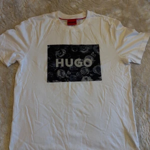 Vit HUGO t-shirt med blommigt tryck - Vit t-shirt från HUGO i bomull med ett stort svart blommigt tryck och HUGO-logga på bröstet. T shirten är i storlek S och helt oanvänd. Köpt men bara legat i garderoben därför skrynklig annars är den helt ny. Hör av vid frågor. 