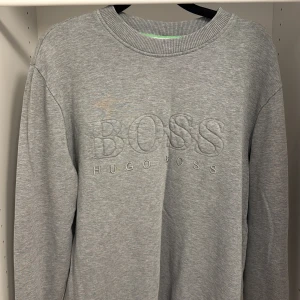 Grå sweatshirt från Hugo Boss - Snygg grå sweatshirt från Hugo Boss med broderad logga framtill. Tröjan har rund halsringning, långa ärmar och en relaxed passform. Perfekt för en stilren och avslappnad look. Tillverkad i mjuk bomullsmix för extra komfort.