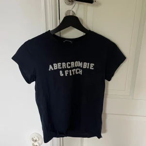 Navy t-shirt från Abercrombie & Fitch - Säljer en svart t-shirt från Abercrombie & Fitch med vit logga tryckt på bröstet. Klassisk rund halsringning och korta ärmar. Perfekt för en avslappnad och stilren look.