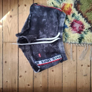 Svarta Levi's shorts med batikmönster och snörning - Säljer ett par svarta shorts från Levi's med batikmönster och vit snörning i midjan. Shortsen har en vit och röd logotypremsa längs kanten och är perfekta för sommaren.
