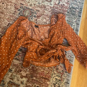 Rostbrun omlottopp med prickar - Trendig rostbrun omlottopp med vita prickar och långa ärmar. Toppen har volangdetaljer vid axlarna och knyts i midjan för en snygg siluett. Perfekt för dig som gillar retrovibbar och vill sticka ut lite extra.