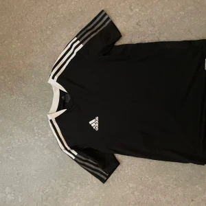 Svart Adidas fotbollströja med vita detaljer - Säljer en svart Adidas fotbollströja med vita ränder på axlarna och vitt runt kragen. Tröjan är kortärmad och tillverkad i lätt, ventilerande material (Primegreen). Adidas-logga på bröstet. Den är använd fåtal gånger och bara luggit      Kom med prisförslag om priset inte passar er😊
