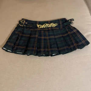 Mörkgrön rutig minikjol med spännen - Säljer en mörkgrön rutig minikjol med plisserad design och två dekorativa spännen i midjan. Kjolen har ett gult broderat tryck framtill och klassiska rutiga linjer i orange och blått. Perfekt för dig som gillar en edgy och trendig stil.