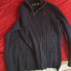 Mörkblå stickad tröja med dragkedja från Polo Ralph Lauren - Säljer en mörkblå stickad tröja från Polo Ralph Lauren med halv dragkedja och klassiskt kabelstickat mönster. Tröjan har lång ärm, hög krage och röd broderad logga på bröstet. Perfekt för en stilren och avslappnad look.
