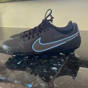 Nike Tiempo Legend fotbollsskor svart/blå - Säljer ett par Nike Tiempo Legend fotbollsskor i svart med blå detaljer och snörning. Skorna har rund tå, platt sula med dobbar och är tillverkade i syntetmaterial. Perfekta för dig som vill ha stilrena och bekväma fotbollsskor på planen.