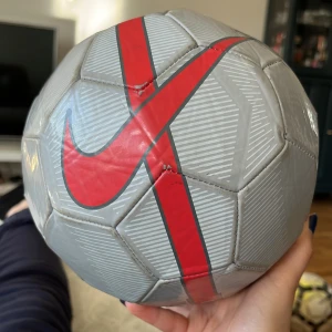 Nike Mercurial Fade fotboll - Säljer en grå Nike Mercurial Fade fotboll med röda och svarta detaljer samt stor Nike-logga. Bollen har ett modernt mönster och är tillverkad i slitstarkt syntetmaterial. Perfekt för matcher eller träning. I storlek 5