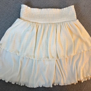 Vit volangkjol med smock - Superfin vit kjol med smock i midjan och dubbla volanger. Kjolen sitter superfint och har ett lite mönstrat tyg. Säljer pga att den är för stor och knappt kommer till användning 💕