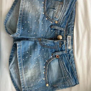 Blå jeansshorts med strass  - Snygga blå jeansshorts med strassdetaljer och slitningar. med hjärtdetalj bak. Perfekta för sommaren, dessutom lågmidjade. Storlek 27! 