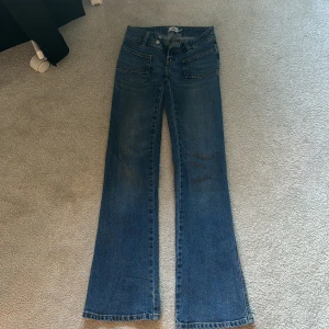 Blå bootcut jeans med dubbla knappar - Säljer ett par blå bootcut jeans med dubbla knappar i midjan och stora framfickor. Jeansen har klassiska bakfickor och är i en klassisk denimtvätt. Perfekta för dig som gillar retrostil och vill ha ett par jeans med lite vidare ben.