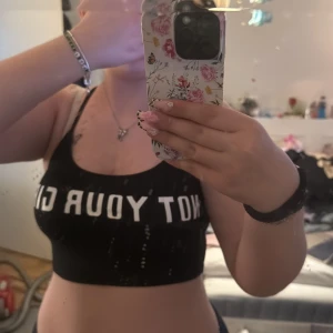 Svart croppad linnetopp från Divided - Svart croppad linnetopp från Divided med smala axelband och vit text 'NOT YOUR GIRL' framtill. Enkel och stilren design, perfekt för en avslappnad look. Tillverkad i bomullsmix för en bekväm känsla.