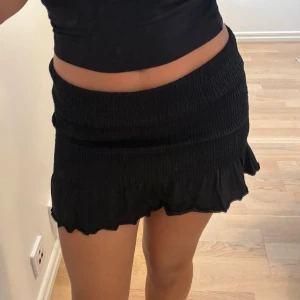 Svart volangkjol - Supersöt svart kortkjol med volangkant och smockad midja. Kjolen har en figurnära passform upptill och faller ut lätt nedtill. Perfekt att styla med en crop top eller t-shirt. Man kan ha den både kort och lång! 