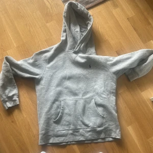 Grå hoodie från Polo Ralph Lauren - Säljer en klassisk grå hoodie från Polo Ralph Lauren med broderad logga på bröstet. Tröjan har huva och en stor magficka framtill. Perfekt för en avslappnad stil och enkel att matcha med det mesta. Den passar barn XL man S och man XS