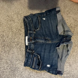 Mörkblå jeansshorts med upprullade ben - Snygga mörkblå jeansshorts från Hollister med upprullade ben och slitna detaljer. Klassisk femficksmodell med knapp och dragkedja framtill. Perfekta för varma dagar och enkel att matcha med olika toppar.