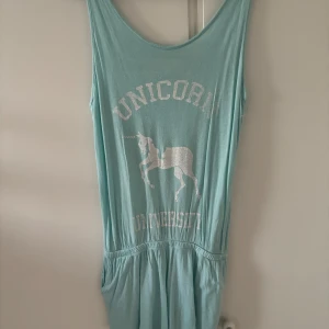 Ljusblå playsuit med unicorn-tryck - Supermjuk ljusblå playsuit med vitt unicorn-tryck och texten 'Unicorn University' framtill. Djup urringning både fram och bak med snygga korsade band i ryggen. Resår i midjan för en bekväm passform.