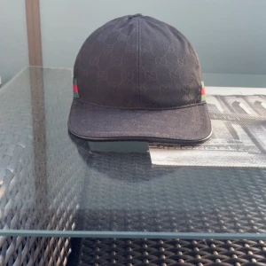 Svart keps från Gucci med GG-mönster - Svart keps från Gucci med diskret GG-monogram över hela tyget och klassiska gröna och röda ränder på sidan. Justerbar rem baktill i svart läder med guldfärgad spänne. Snygg och stilren accessoar.