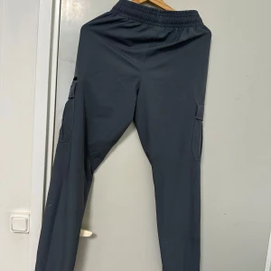 Mörkgrå cargobyxor från Under Armour - Säljer ett par mörkgrå cargobyxor från Under Armour med elastisk midja och dragsko. Byxorna har flera fickor på sidorna och en smalare passform. Perfekta för träning eller chill.