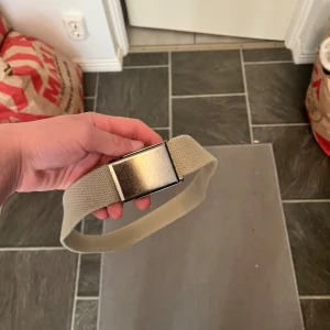 Grisch bälte - Säljer ett stilrent beige bälte i tyg med ett rektangulärt silverfärgat metallspänne. Perfekt för att ge en avslappnad touch till din outfit. Justerbar längd och enkel att använda.