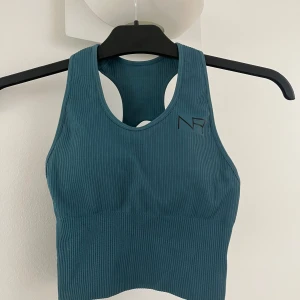 Blå ribbad sporttopp från NA-KD - Säljer en blå ribbad sporttopp från NA-KD med brottarrygg och svarta spännen baktill. Toppen har en tight passform och är perfekt för träning. Snygg och enkel design med diskret logga framtill. Andvänd FÅTAL gånger, vet ej storleken då jag tydligen har klippt av lappen men passar på mig som är xs/s och har större bröst. Den är ganska stretchig så tror den skulle passa även större storlekar!