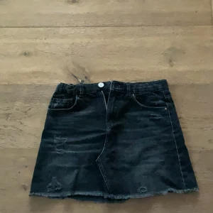Svart jeanskjol från Zara - Svart jeanskjol från Zara med rå kant nertill och slitna detaljer. Klassisk femficksmodell med knapp och dragkedja framtill. Perfekt för en cool och avslappnad stil.