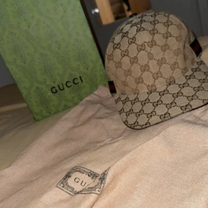 Beige keps från Gucci med GG-mönster - Säljer en beige keps från Gucci med det klassiska GG-monogrammet över hela kepsen. Kepsen har bruna detaljer och en röd-grön rand på sidan. Märkt med storlek L (59 cm) och har insida i brunt tyg samt läderdetaljer.