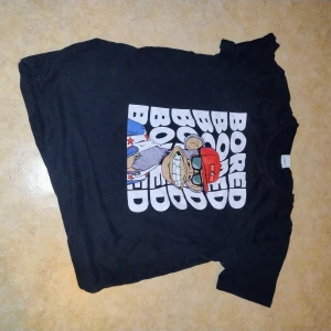 Svart t-shirt med apa-tryck och text - Cool svart t-shirt med stort tryck av en tecknad apa med röd keps och texten 'BORED' upprepad flera gånger på framsidan. T-shirten har rund hals och korta ärmar. Perfekt för dig som gillar streetwear och unika motiv.