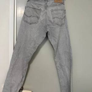 Ljusgrå jeansbyxor från Levis - Ljusgrå jeansbyxor från Levis med klassisk femficksdesign och raka ben. Byxorna har Levis-logga på bakfickan och läderpatch i midjan. Perfekta för en avslappnad stil.