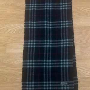 Rutig halsduk från Burberrys - Säljer en klassisk rutig halsduk från Burberrys i mörka färger med inslag av rött och vitt. Halsduken har fransar i ändarna och är tillverkad i mjuk ull. Perfekt accessoar för kyliga dagar.