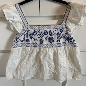 Vit och blå broderad linnetopp - Söt vit linnetopp med blått broderi framtill och volangärmar. Toppen har breda axelband med blått rutmönster och knytning i ryggen. Perfekt för varma dagar och ger en romantisk känsla.