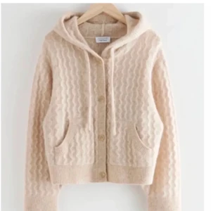 Beige stickad hoodie med knappar - EFTERTRAKTAD Mysig beige stickad hoodie från And Other Stories med vågigt mönster, knappar framtill och två fickor. Tröjan har långa ärmar, ribbade muddar och huva med dragsko. Perfekt för lager-på-lager och chill dagar.