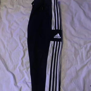 Svarta Adidas träningsbyxor med vita ränder - Säljer ett par svarta Adidas träningsbyxor med klassiska vita ränder längs sidorna och Adidas-logga på benen. Byxorna har resår i midjan och är tillverkade i ett mjukt och bekvämt material, perfekta för sport eller chill.