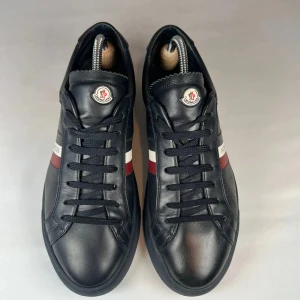 Svarta sneakers från Moncler med röd och vit detalj - Snygga svarta sneakers från Moncler med klassisk design. Skorna har svarta snören, rund tå och en röd samt vit rand på sidan med Moncler-logga på plösen. Tillverkade i skinn och passar perfekt till en stilren vardagslook.
