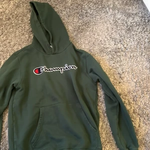 Mörkgrön hoodie från Champion - Säljer en mörkgrön hoodie från Champion i storlek 2X-Large. Tröjan har klassisk huva, magficka och stort broderat Champion-logo på bröstet. Perfekt för en avslappnad stil och enkel att matcha med jeans eller joggers.