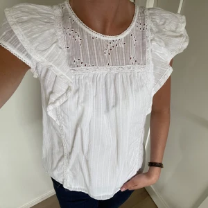 Vit blus med volang -  Söt vit blus med broderade spetsdetaljer upptill och volang. Blusen har korta ärmar och är perfekt nu till sommaren!