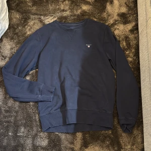 Mörkblå sweatshirt från GANT - Säljer en stilren mörkblå sweatshirt från GANT med broderad logga på bröstet. Tröjan har rund halsringning, långa ärmar och ribbade muddar vid ärmslut och nederkant. Perfekt för en avslappnad och klassisk look.