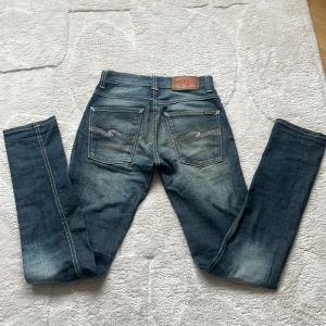 Blå jeans från Nudie Jeans - Säljer ett par klassiska blå jeans från Nudie Jeans med raka ben och snygga slitningar. Jeansen har knappar i gylfen och detaljerade bakfickor med Nudie-loggan. Perfekta för dig som gillar en tidlös denimlook.