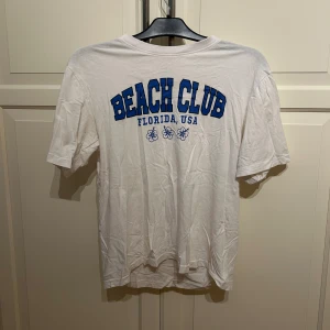 Vit t-shirt med blå BEACH CLUB-tryck från 157 - Vit over sized t-shirt från 157 med blå text 'BEACH CLUB FLORIDA, USA' och små blommor framtill. Klassisk passform och rund halsringning. Tillverkad i 100% bomull, i strl S