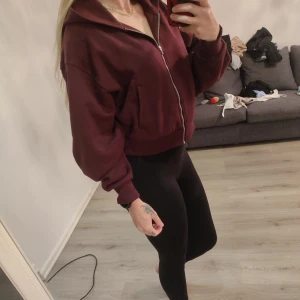 Vinröd zip-hoodie - Mysig vinröd hoodie med dragkedja och huva. Modellen är croppad och har långa ärmar samt breda muddar vid ärmslut och nederkant. Perfekt för en avslappnad stil.