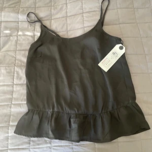 Svart linnetopp med volang från ONLY - Säljer en svart linnetopp från ONLY med smala axelband och volang nertill. Toppen har omlottdetalj i ryggen och justerbara axelband. Perfekt för varma dagar eller att styla lager på lager.