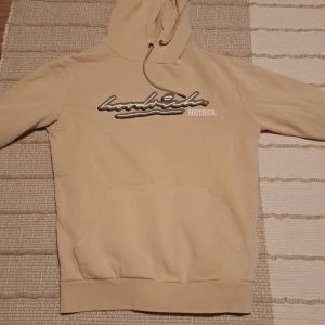 Beige hoodie från Hoodrich - Säljer en beige hoodie från Hoodrich med svart och vit logga broderad på bröstet. Tröjan har huva med dragsko och en stor magficka framtill. Perfekt för en avslappnad stil.