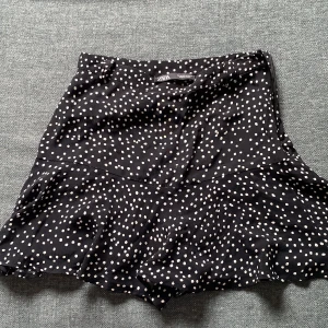 Svart prickig kjol från Zara - Supersöt svart kjol från Zara med vita prickar och volang nertill. Kjolen har en kort passform och är tillverkad i ett lätt och mjukt polyestertyg. Perfekt för dig som gillar lekfulla mönster och vill ha en kjol som sticker ut.